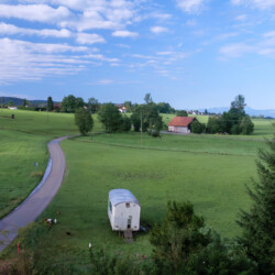 Ausblick vom Balkon Sommerliche Landschaft mit grünen Wiesen, blauem Himmel, einer Straße die sich von vorne nach hinten schlängelt und im Hintergrund Berge.